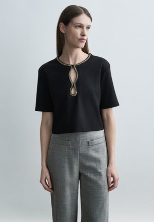 Žena nosi crnu majicu kratkih rukava s zlatnim ukrasnim perlicama na izrezu i izrezu, uparenu s visokim sivim teksturiranim hlačama.