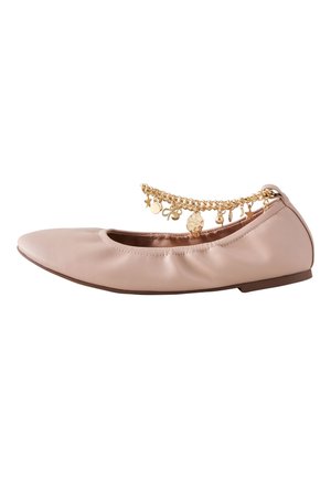 Jambes portant des ballerines roses et un bracelet de cheville doré avec un charme, debout sur un sol clair, avec une jupe plissée marron au-dessus des chevilles.