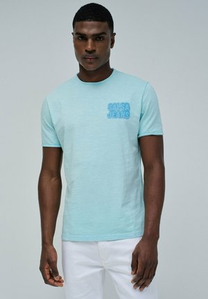Camiseta de manga corta de color azul claro con un acabado texturizado y un parche del logo "SALSA JEANS" en el lado izquierdo del pecho. Combinada con pantalones blancos.