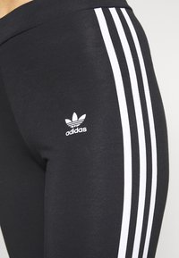 Černé legíny vyrobené z elastického materiálu se třemi bílými pruhy a malým bílým logem Adidas blízko kyčle.