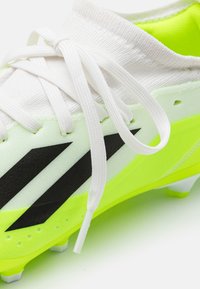 Botas de fútbol blancas con acentos en amarillo neón. Cuentan con un upper sintético suave, cordones tonales y un diseño de franjas negras en el lateral.