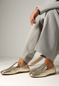 Metalické zlaté slip-on boty se zaoblenými perforacemi, plochou podrážkou a měkkou, hladkou texturou. V kombinaci se světle šedými širokými kalhotami.