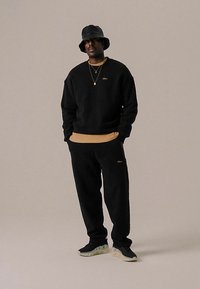 Sweatshirt en polaire noir et pantalon de survêtement superposés à un haut beige. Porté avec un chapeau seau noir et des baskets avec une semelle claire.