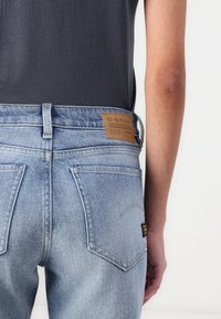 Světle modré džíny z denimu s hnědým koženým štítkem označeným "G-Star Originals", mají design s pěti kapsami a mírné vyšisování na zadní straně.