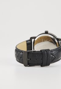 Black leather watch strap with a textured finish, featuring a silver-tone buckle and soft beige lining. Round watch face visible.