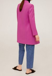 Pitkä vaaleanpunainen takki suoralinjaisella leikkauksella, sivutaskuilla ja lovitaskolla, yhdistettynä rentoihin sinisiin farkkuihin ja mustiin liukuhihna-sandaaleihin.