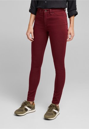 Osoba ubrana w dopasowane burgundowe spodnie, czarną koszulę z podwiniętymi rękawami i zielone trampki z białymi i brązowymi akcentami, stojąca na szarym tle.