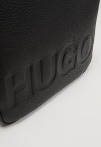 Schwarze Lederhandtasche mit strukturierter Oberfläche, versehen mit eingeprägtem "HUGO"-Logo. Rundliche Form mit klaren Kanten und ohne sichtbare Hardware.