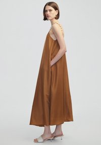 Vestido maxi marrón sin mangas con bolsillos laterales, confeccionado en una tela suave. Presenta tirantes ajustables de trenzado. El modelo lleva sandalias deslizantes blancas.
