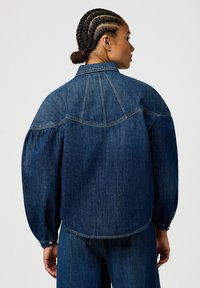 Veste en denim avec une coupe décontractée, des manches bouffantes et un col classique. Présente une teinte bleue, des coutures subtiles et des poignets boutonnés.