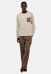 Chemise beige à manches longues avec une poche plaquée marron, associée à un pantalon marron ample. Matériau en coton avec une texture lisse.