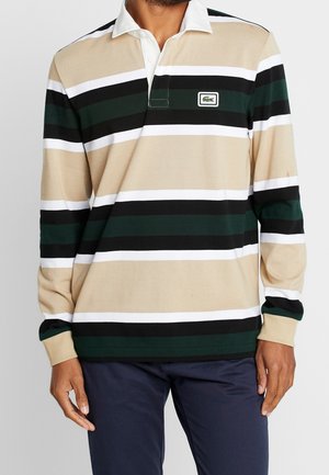 Mężczyzna ubrany w beżową, białą, czarną i ciemnozieloną poziomo paskowaną koszulę polo z długim rękawem, z białym kołnierzykiem i granatowymi spodniami.