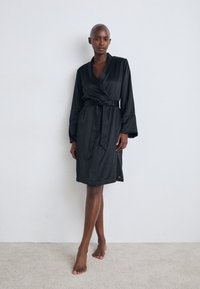 Svart satinbadrock med omslagdesign, långa ärmar och bälte. Slät textur och knälång klippning med en sidorslits.