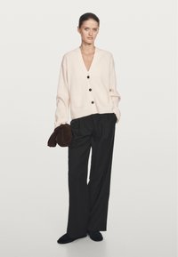 Licht crèmekleurige v-hals cardigan met drie knopen, twee voorkruiszakken, gedragen over zwarte wijde broek. Model houdt een donkerbruine accessoire vast.