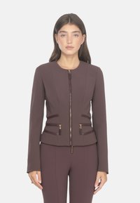 Giacca marrone con zip, dal design aderente, con cerniere a contrasto, maniche lunghe e cuciture strutturate. Si abbina a pantaloni marroni coordinati.