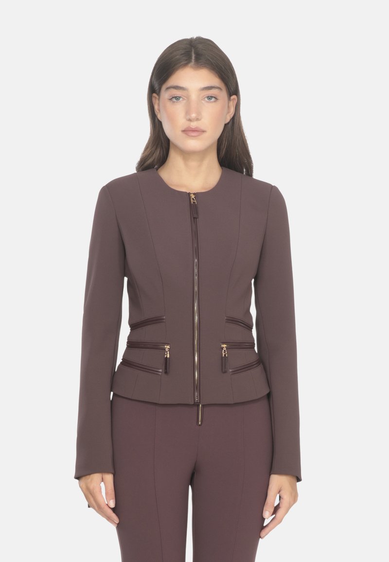 Giacca marrone con zip, dal design aderente, con cerniere a contrasto, maniche lunghe e cuciture strutturate. Si abbina a pantaloni marroni coordinati.