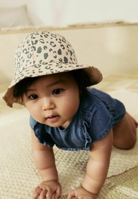 Un chapeau à larges bords en imprimé léopard, une combinaison en denim avec des manches volantes, sur un bébé qui rampe. Surface texturée beige doux en arrière-plan.