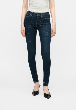 Femme portant un jean skinny foncé avec de petites taches blanches, un top blanc sans manches et des chaussures pointues noires avec des lanières.
