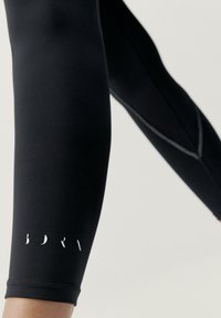 Leggings neri con una texture liscia, dotati di un logo bianco vicino all'orlo. Il design include bordi senza cuciture e una silhoutte aderente.