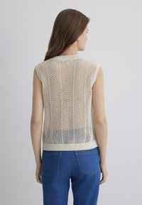 Ärmelloser Strickpullover in Creme mit einem Zickzack-Ölmuster; verfügt über einen gerippten Halsausschnitt und Saum, kombiniert mit blauem, hochgeschnittenem Jeans.