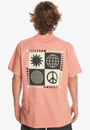 Person mit lockigem braunem Haar trägt ein rosa T-Shirt mit einem schwarz-weißen Quadratdesign auf dem Rücken, das Sonne, Globus, Spirale und Friedenssymbole zeigt.