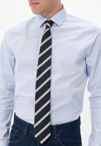 Camisa de vestir a rayas azul claro con cuello clásico, combinada con una corbata azul marino que presenta rayas diagonales beige y blancas. Llevada con jeans de mezclilla oscuros.