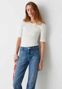 Witte geribbelde top met korte mouwen gecombineerd met hoogopgetrokken blauwe spijkerbroek. De outfit heeft een subtiele textuur en wordt geaccentueerd met een gemusterde sjaal.