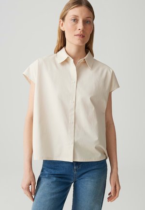 Femme debout portant une chemise crème à manches courtes boutonnée et un jean bleu sur un fond neutre uni.