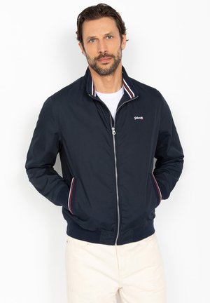 Mężczyzna ubrany w granatową kurtkę na zamek z paskiem w paski na kołnierzu i wykończeniach kieszeni, białą podkoszulkę oraz beżowe spodnie, stojący z rękami w kieszeniach.