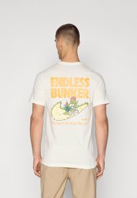 Camiseta blanca de algodón con un gráfico colorido de un personaje descansando y el texto "BUNKER INFINITO" y "La búsqueda de cualquier otra mentira."