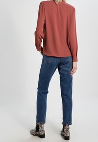 Blouse couleur rouille à manches longues, associée à un jean bleu délavé. Le chaussage présente un motif léopard noir et blanc.