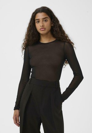 Junge Frau mit langen lockigen Haaren, die ein enges, durchsichtiges schwarzes Langarmoberteil und schwarze High-Waist-Hosen trägt, steht mit einer Hand in der Tasche.