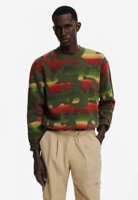 Žalios kamufliažinės spalvos megztinis su raudonomis ir geltonomis detalėmis, su pūstais apvaliais kaklo kraštais ir tekstūruotu mezginiu. Dėvimas su smėlio spalvos kelnėmis.