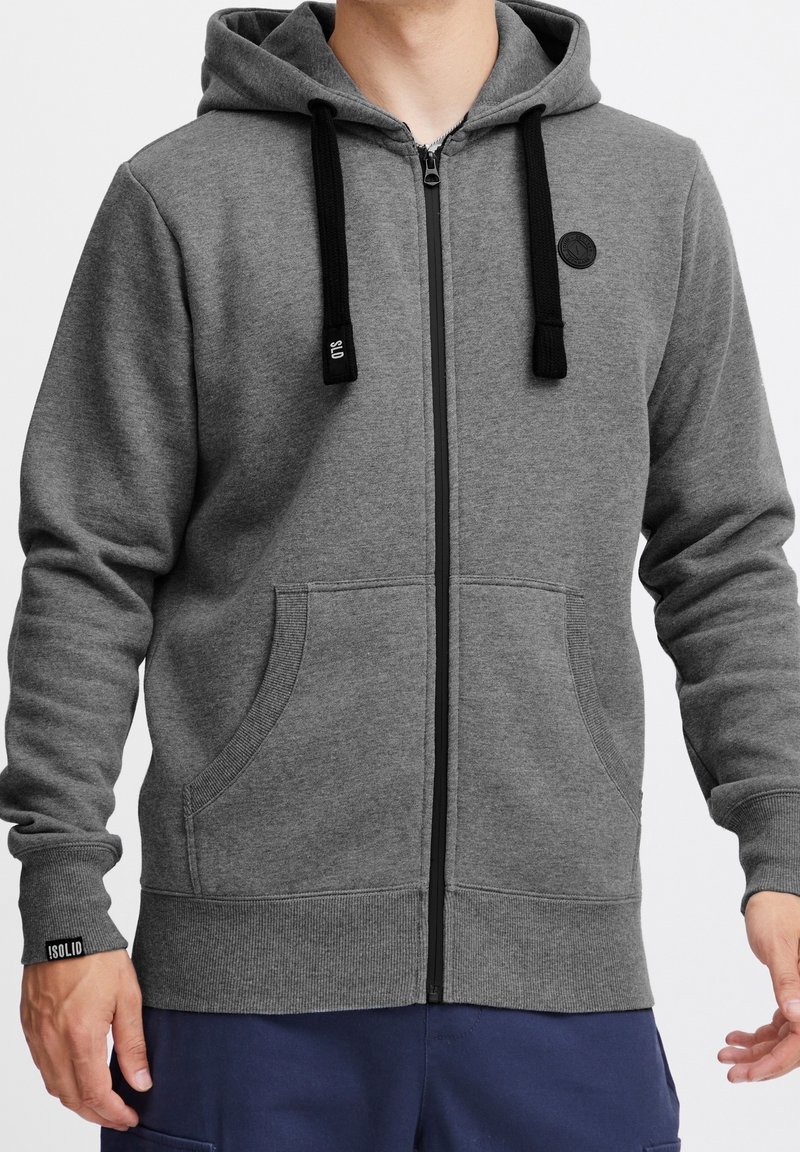 Sudadera gris con cremallera y cordones negros, que cuenta con un parche de logo, dos bolsillos frontales y puños y dobladillo acanalados.