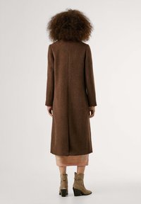 Long brown woolen coat with a straight cut and single seam down the back; worn over a pink knitted skirt and paired with beige heeled boots.