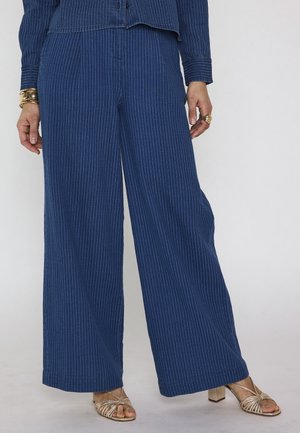 Vrouw draagt blauwe broek met wijde pijpen en krijtstrepen, bijpassende top, gouden armbanden en ringen, en gouden sandalen met hak en bandjes.