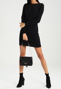 Robe en tricot noire à manches longues et décolleté en dentelle en V, accompagnée d'un sac noir avec une bandoulière en chaîne et des accents texturés.