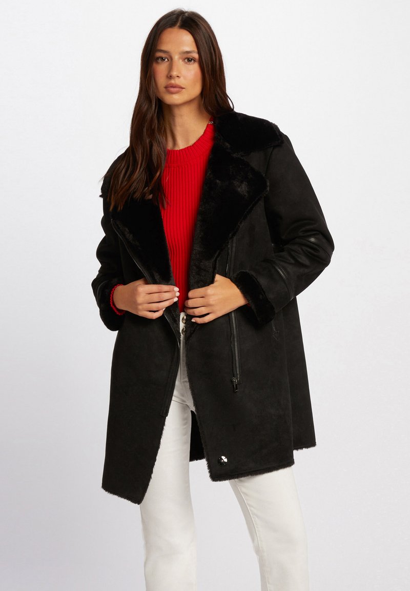 Svart faux fur-foret jakke med bred krage, glidelås og sidelommer. Brukt over en rød ribbestrikket genser og kremfargede jeans.