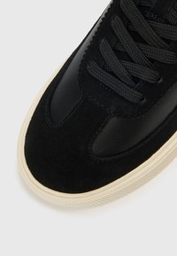 Ténis preto com combinação de materiais em camurça e couro, com atacadores planos pretos e sola texturizada de cor creme.