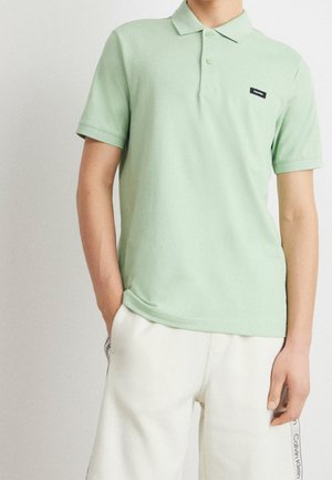 Personne portant un polo vert clair avec un petit logo et un short blanc avec la marque Calvin Klein sur le côté.