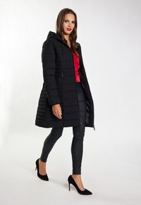 Parka nera con cappuccio, dotata di chiusura zip e tasche laterali. Indossata sopra una maglietta rossa, abbinata a pantaloni skinny neri e tacchi.