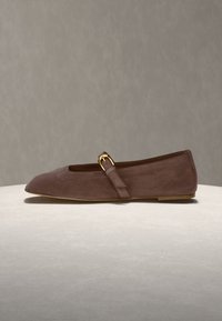 Chaussure plate en daim marron avec bout pointu et une bride unique avec une boucle dorée, présentée sur une surface claire.