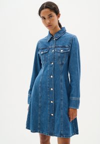 Robe en denim à manches longues, col classique, fermeture à boutons sur le devant et poches poitrine. Couleur bleue avec des détails de couture visibles.