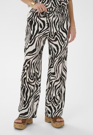 Kvinde iført højttaljede bukser med vide ben i sort-hvide zebra-striber, kombineret med brune kork-platformsandaler og guldsmykker.