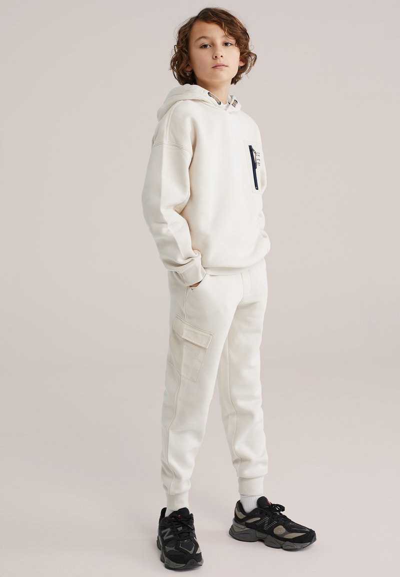 Jonge persoon met krullend haar staat en draagt een gebroken witte hoodie, bijpassende cargobroek joggers, witte sokken en zwarte sportieve sneakers.