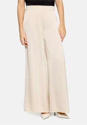 Vrouw die beige hoog getailleerde, wijd uitlopende broek draagt, gecombineerd met witte puntige hakken en een ingestopt zwart topje.