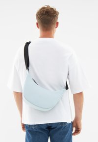 Borsa a spalla di colore azzurro chiaro con design curvo, tracolla nera regolabile e cerniera; texture liscia con hardware minimal.