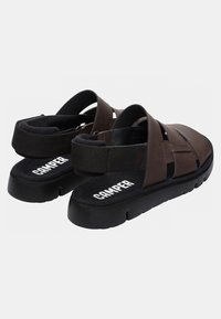 Brown leather sandals with a black rubber sole and adjustable strap. Features a textured insole and branding on the footbed.