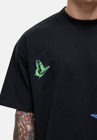 T-shirt in cotone nero con farfalla verde e dettagli blu sulla parte anteriore. Design con collo tondo, maniche corte e vestibilità rilassata.