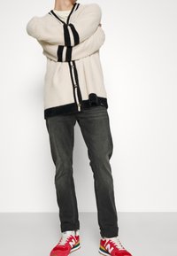 Texturovaný slonovinový cardigan s černými pruhy, s knoflíkovým zapínáním, zkombinovaný s tmavými džíny a červenými teniskami s vícebarevnými akcenty.